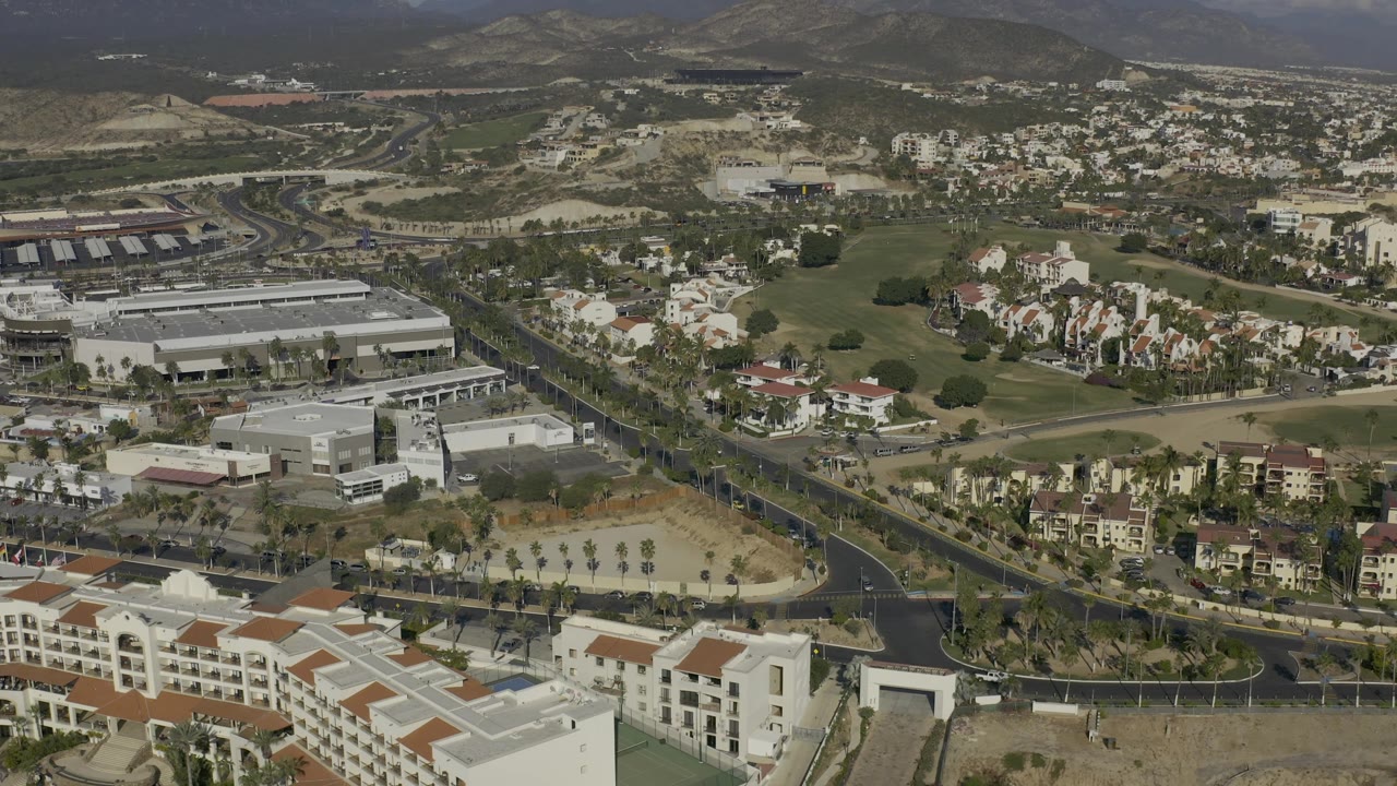 Mid-clip aerial view over Baja California Sur with foreground white resort-style buildings, a dark road junction, scattered homes, landscaped green strips, mountains, and no visible people.