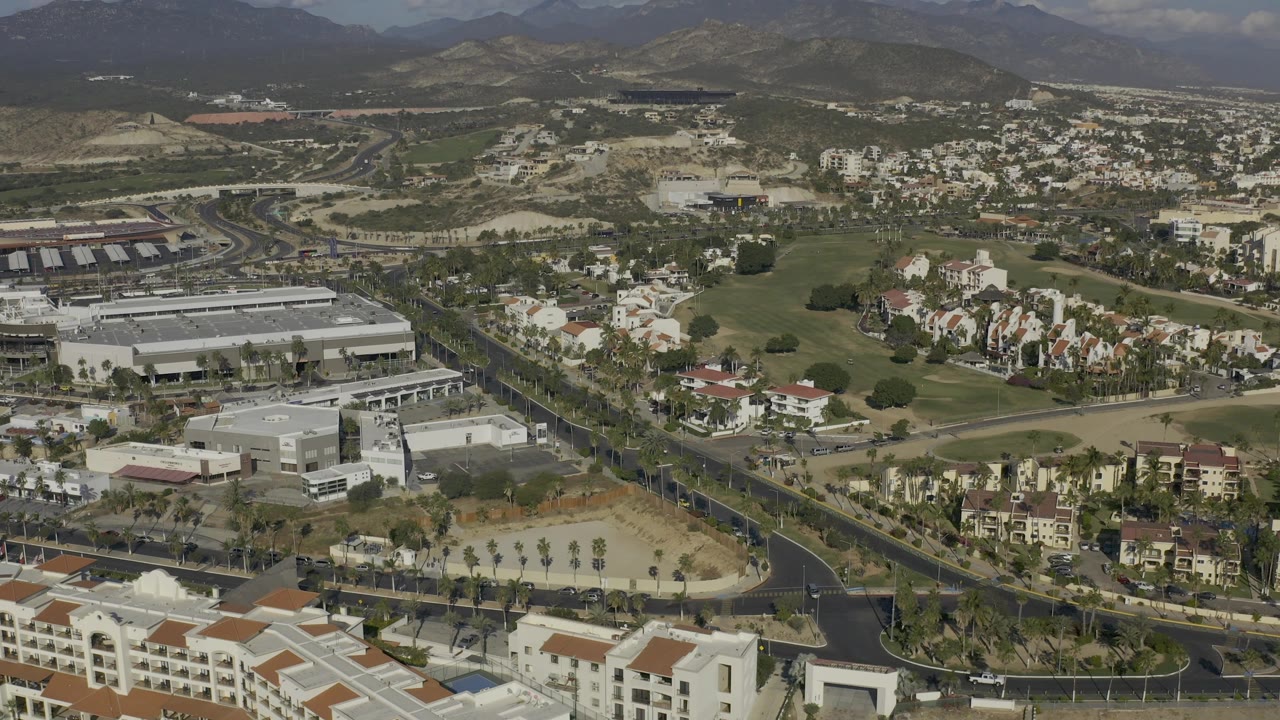 Aerial town view with a broad road, large buildings, houses, palms, and hills.