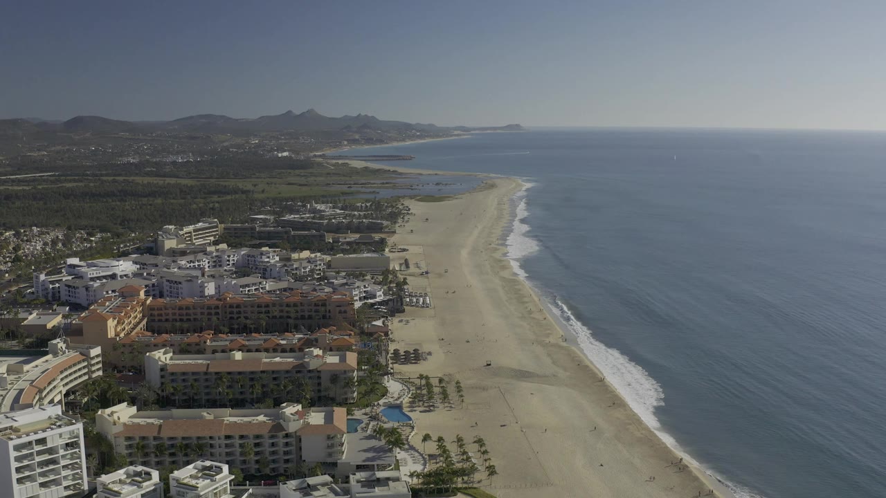 High aerial view in Baja California Sur, Mexico looking along the beach as the surf line curves past large coastal buildings and small figures on the sand.