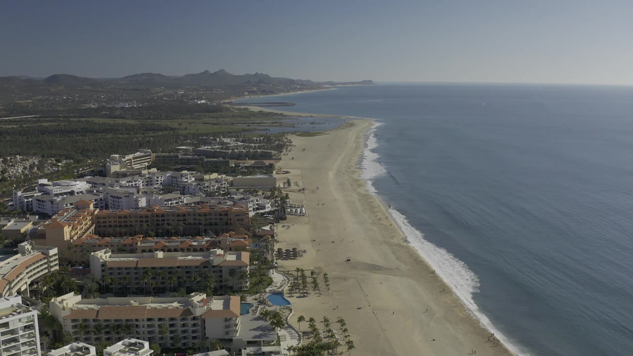 Aerial coastline view with beachfront buildings, sand, surf, and hills.