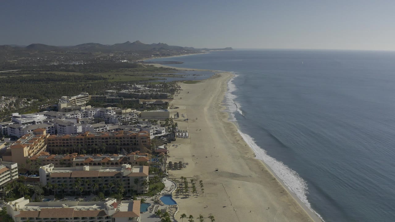 Opening aerial frame over Baja California Sur, Mexico with beachfront resort buildings at lower left, a wide pale sand beach, blue ocean to the right, distant hills, and no visible people.