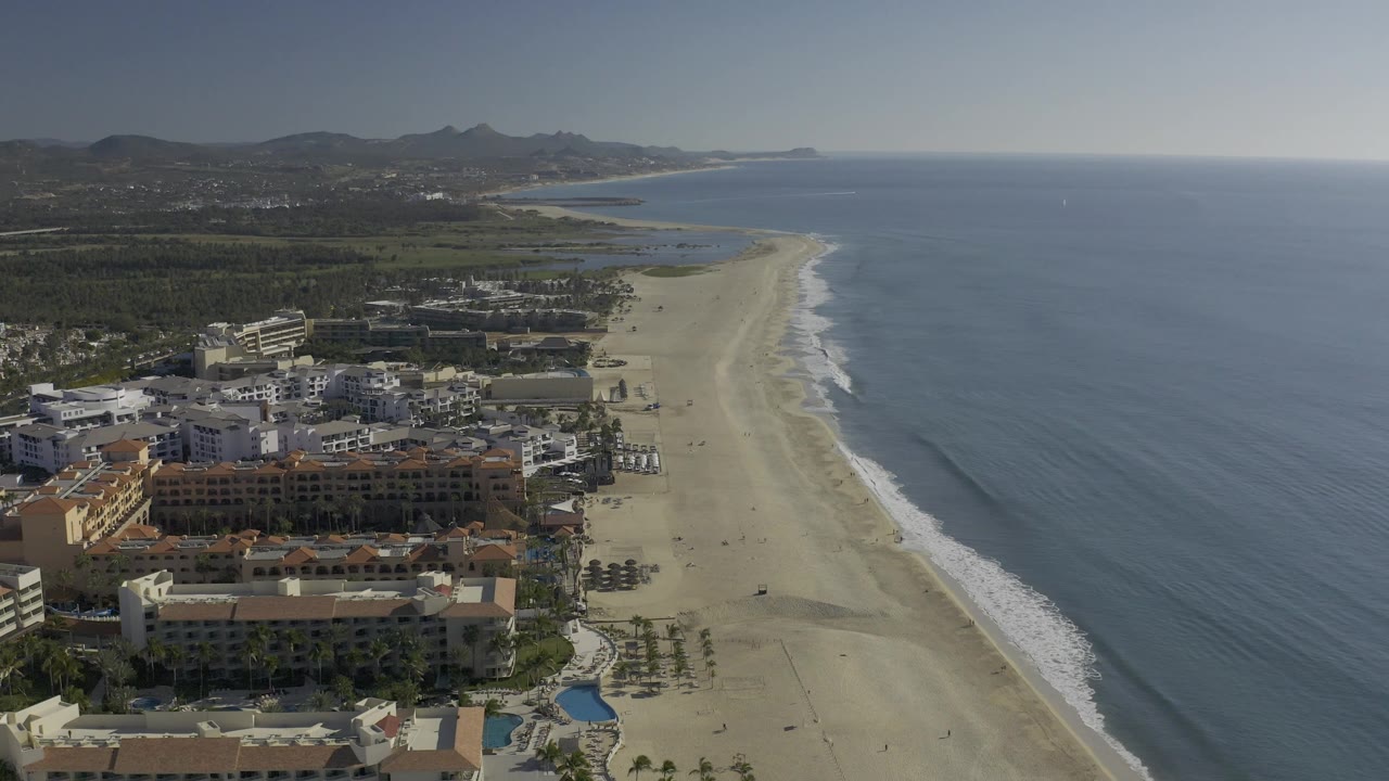 Baja California Sur aerial frame showing resort roofs in the foreground, a long clean beach curving toward distant hills, blue water at right, and no visible people.
