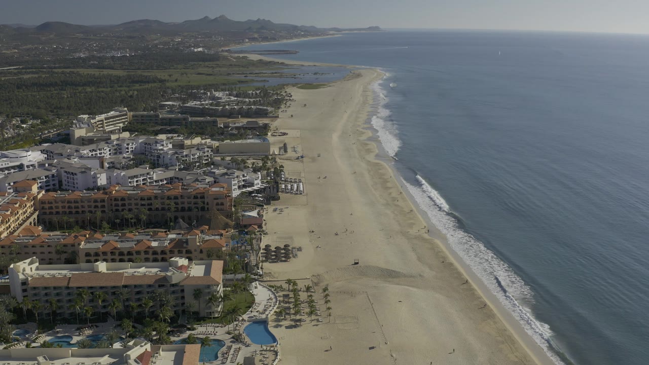 Aerial view over Baja California Sur beach with resort-style buildings, pools, palm rows, surf lines, open blue water, distant hills, and no visible people.