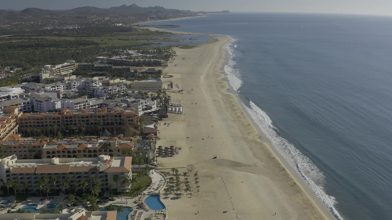 Drone-style view of Baja California Sur shoreline with a broad sandy beach, white surf bands, beachfront structures, pools, palms, open water to the right, and no visible people.