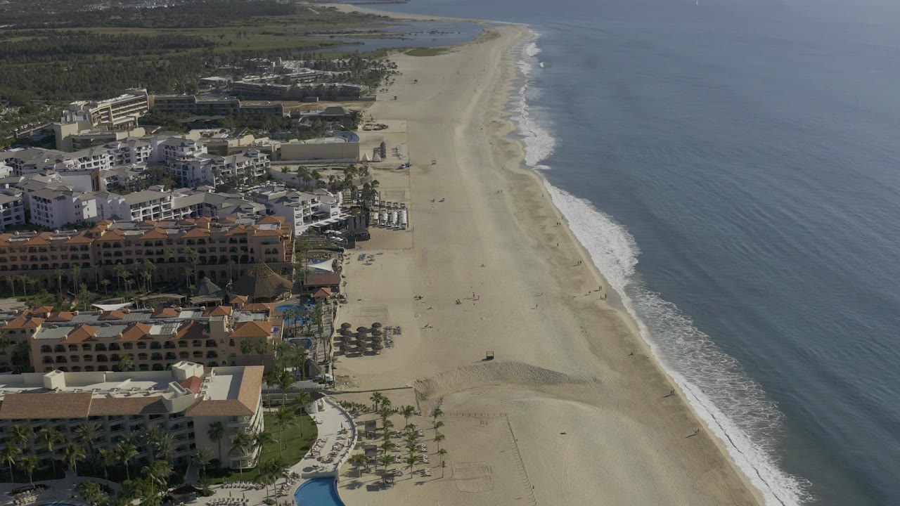 Elevated aerial frame over Baja California Sur, Mexico, with beachfront buildings on the left, a wide sand beach down the center, blue ocean on the right, and no visible people.