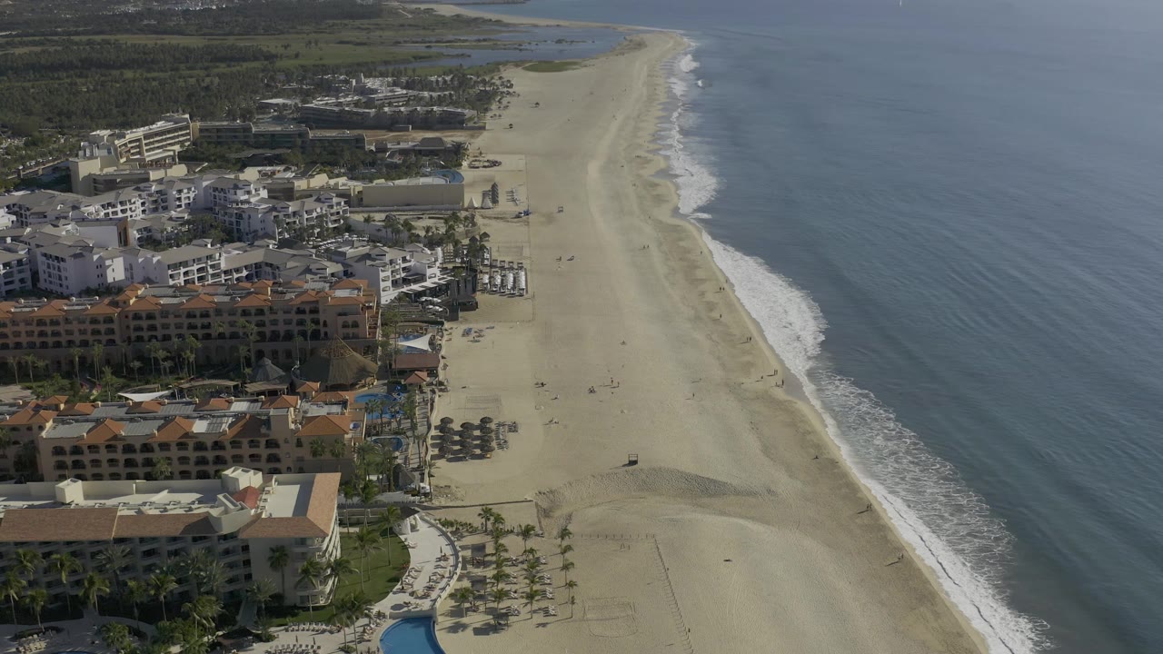 High coastal view with resort-style buildings, beach umbrellas, surf, and open sea.