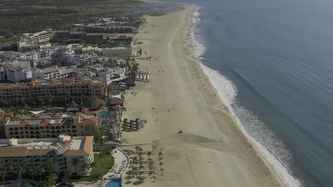High aerial frame over Baja California Sur, Mexico, with beachfront development clustered left of the sand, a pale beach strip, rippled blue water, and no visible people.