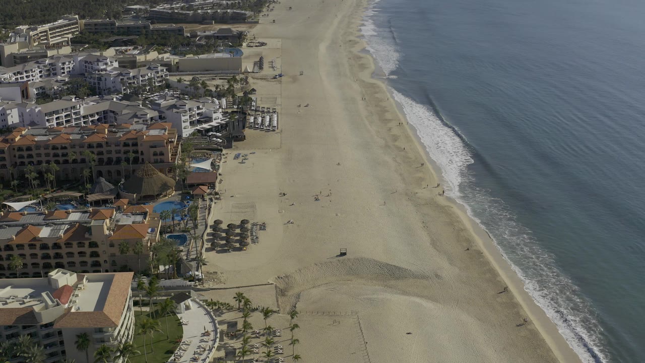 Aerial frame over Baja California Sur, Mexico showing beachfront buildings at left, a wide sand beach in the middle, blue ocean at right, and no recognizable people.