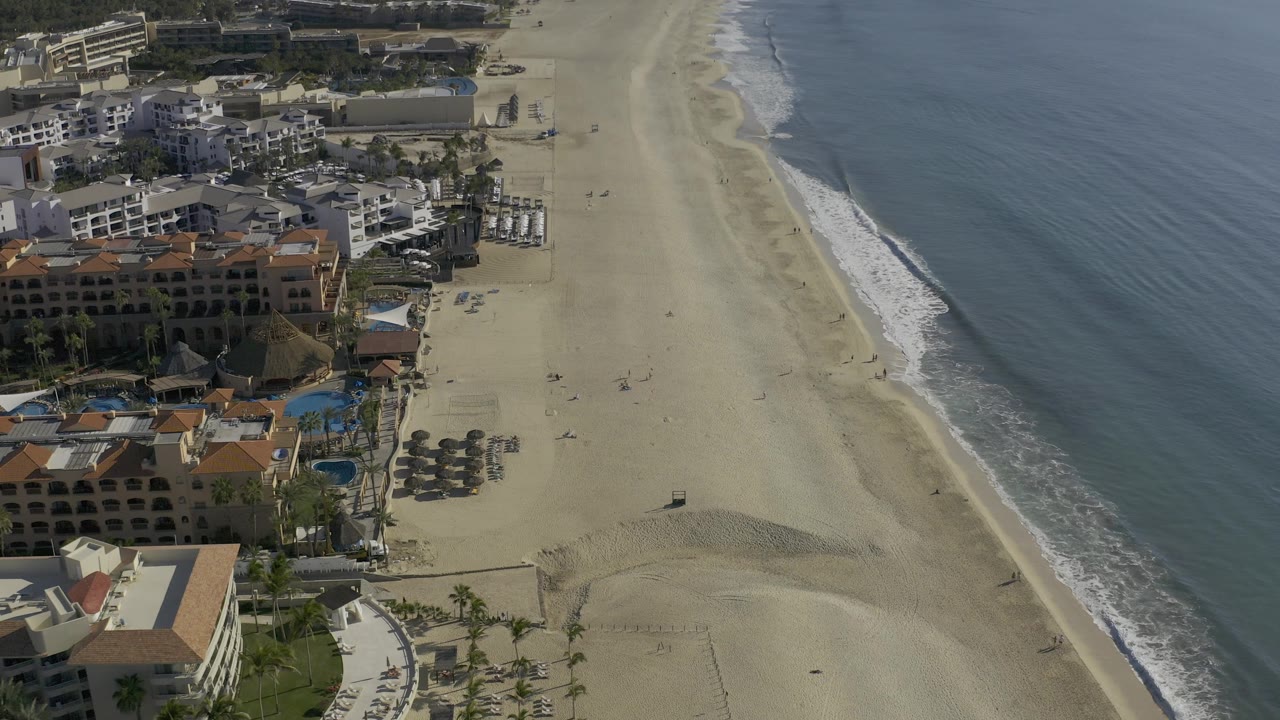 Baja California Sur, Mexico aerial frame showing resort-like buildings with red roofs, a broad empty-looking beach, small wave bands, and no recognizable people.