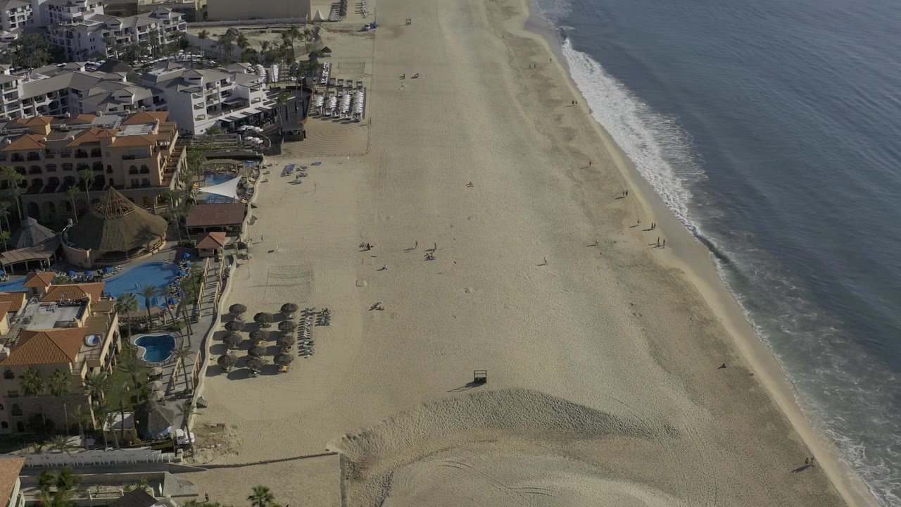 Mid-clip aerial view over Baja California Sur beach, with the ocean occupying the right side and clustered coastal buildings and blue pools along the left edge.