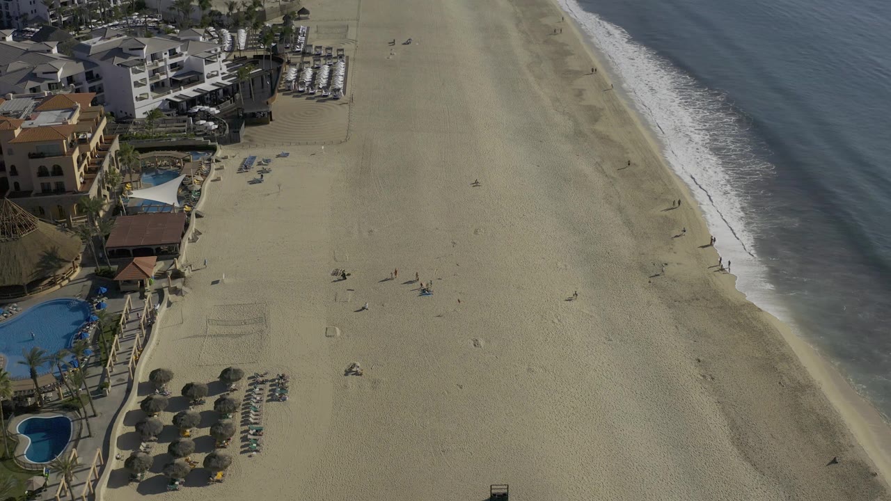 Aerial view one second in over Baja California Sur, showing more open sand between the resort-side pools and the diagonal surf line, with a few tiny distant figures on the beach.