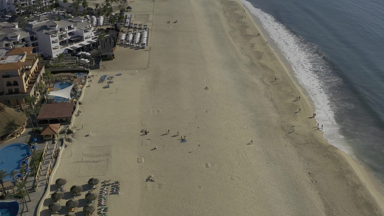 Mid-clip aerial frame over Baja California Sur beach, with the surf line tighter at the right edge, rows of loungers near the lower left, pools and buildings above them, and no recognizable people.