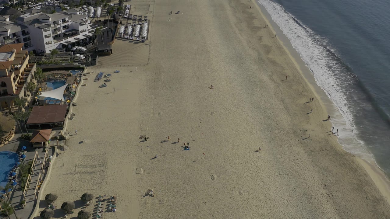Aerial coastline with open beach, diagonal surf, buildings, and pool areas.