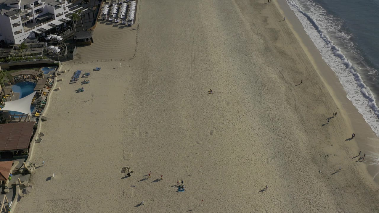 Aerial frame over a Baja California Sur beach with resort buildings at left, pale sand across the center, surf along the right edge, and tiny distant figures with no readable faces.
