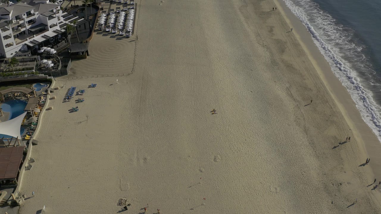 Drone frame over Baja California Sur showing resort roofs near the upper left, orderly rows of beach chairs, open sand, and white surf running along the right side with no recognizable faces.