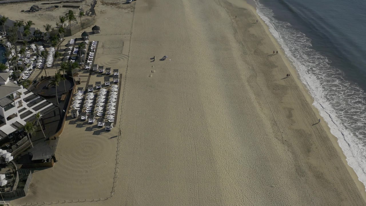 A Baja California Sur shoreline from above, showing white umbrellas and loungers along the left edge, a wide central sand strip, a narrow surf line, and no visible people.