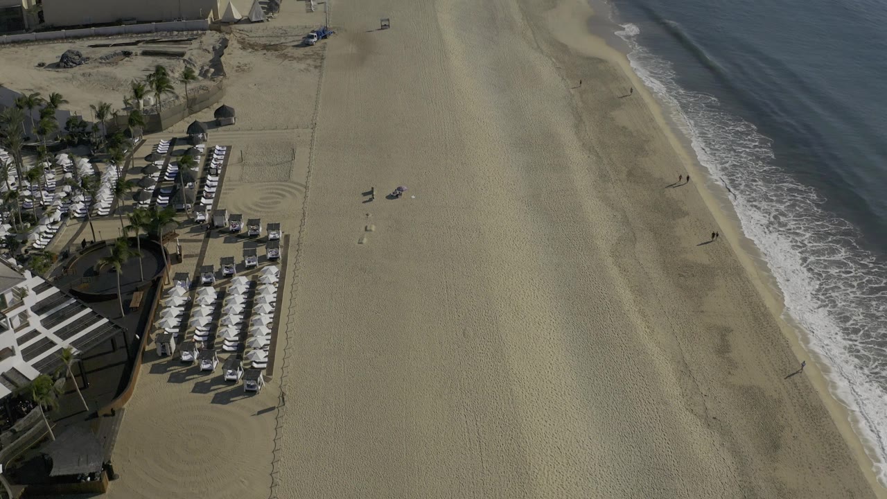 A high oblique view of Baja California Sur coast with the umbrella seating area shifted lower left, open raked sand across the frame, blue water to the right, and no visible people.