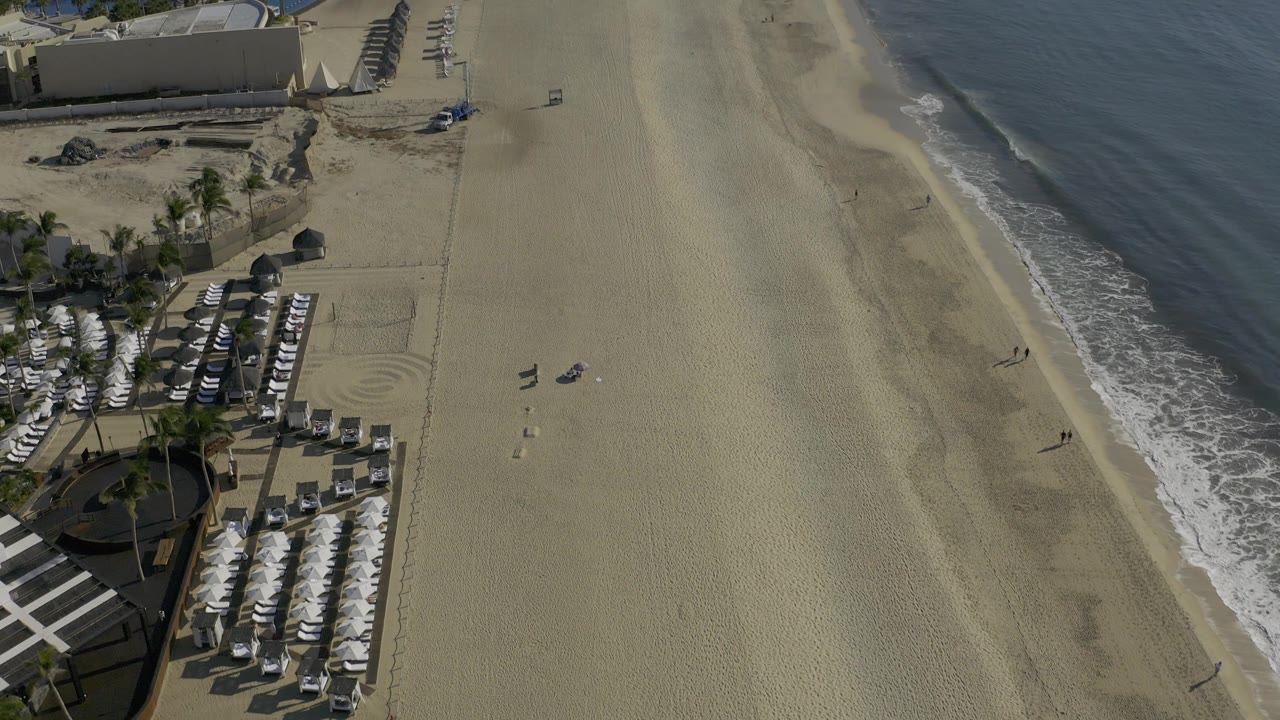 A Baja California Sur beach frame with the umbrella rows lower left, a long uninterrupted tan sand area through center frame, the ocean along the right edge, and no visible people.