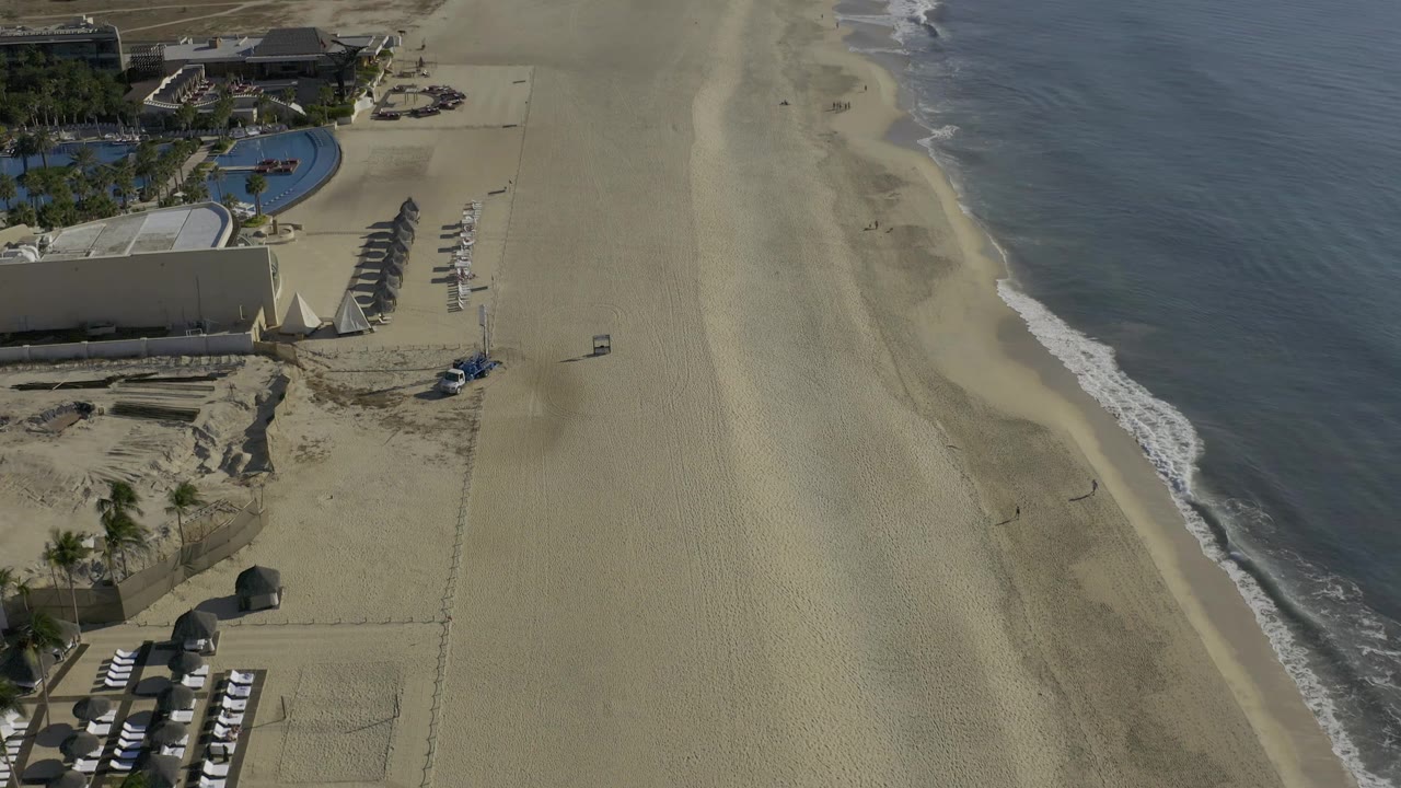 High aerial frame of the Baja California Sur shoreline, with the pool and shade structures on the left, a small vehicle near the center sand, soft surf on the right, and no visible people.