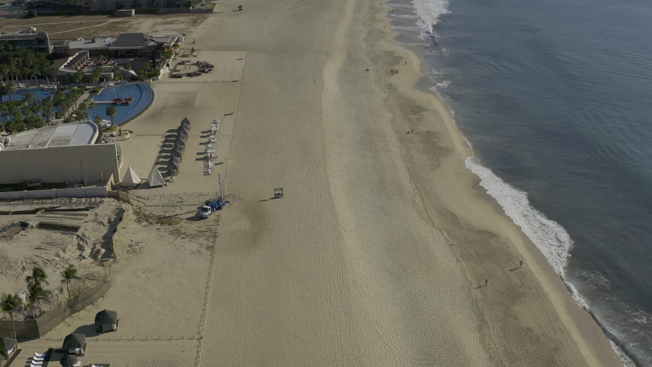 Aerial view along the Baja California Sur coast with beachfront buildings and pool at upper left, open sand through the middle, a narrow surf band, and no visible people.