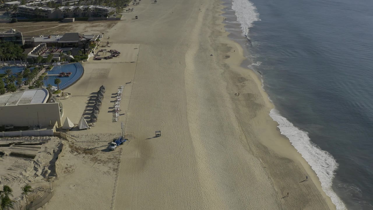 Drone-height frame over Baja California Sur showing a curved pool, rows of shade structures or loungers, broad empty sand, a small vehicle, and waves breaking along the right edge with no visible people.
