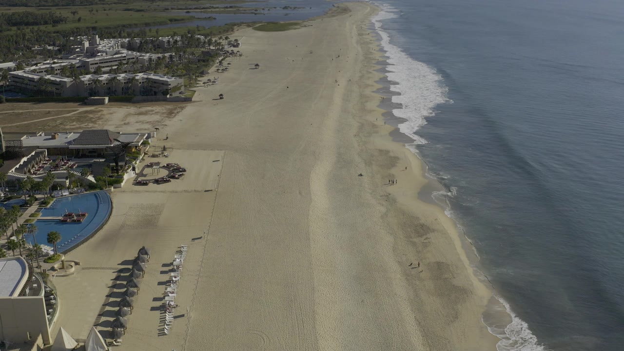Aerial frame over Baja California Sur, Mexico, showing a long sandy beach, calm blue ocean, low resort buildings, pool areas, inland wetlands, and tiny people on the sand.
