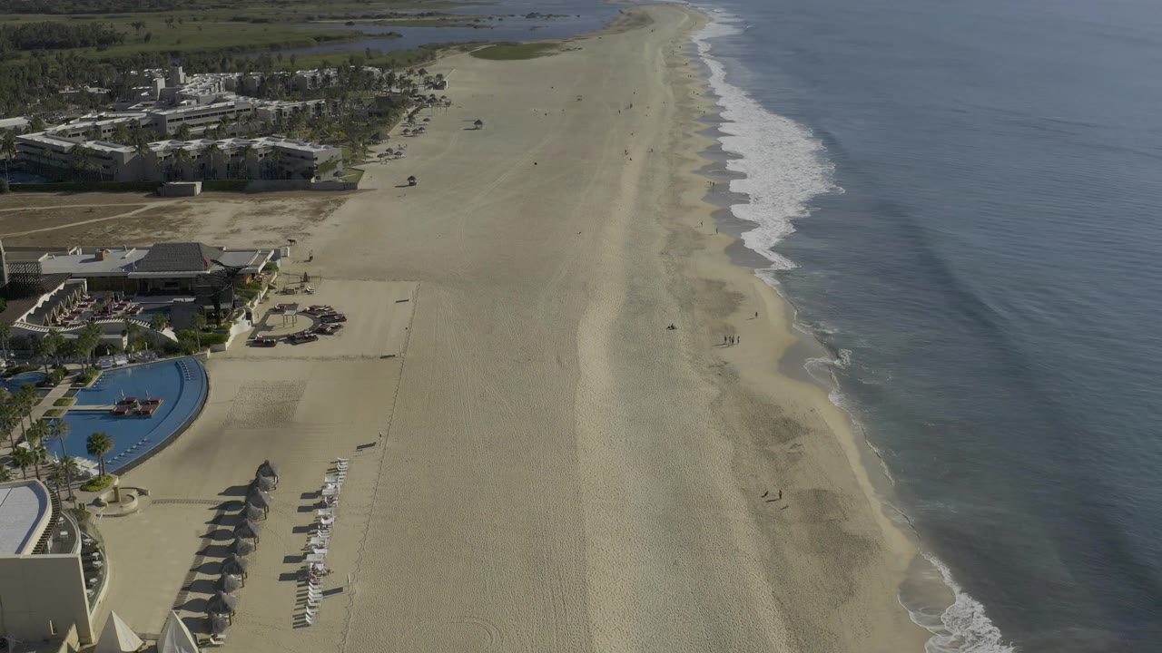 Drone view down a broad beach with surf lines and resort grounds at the edge.
