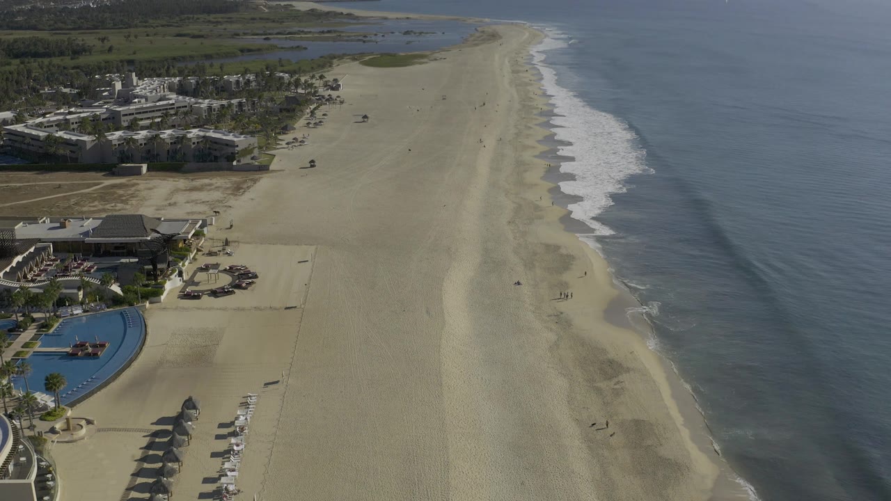 High aerial frame over Baja California Sur, Mexico, with resort buildings at lower left, pale sand beach through center, low waves on the right, wetlands inland, and tiny figures on the beach.