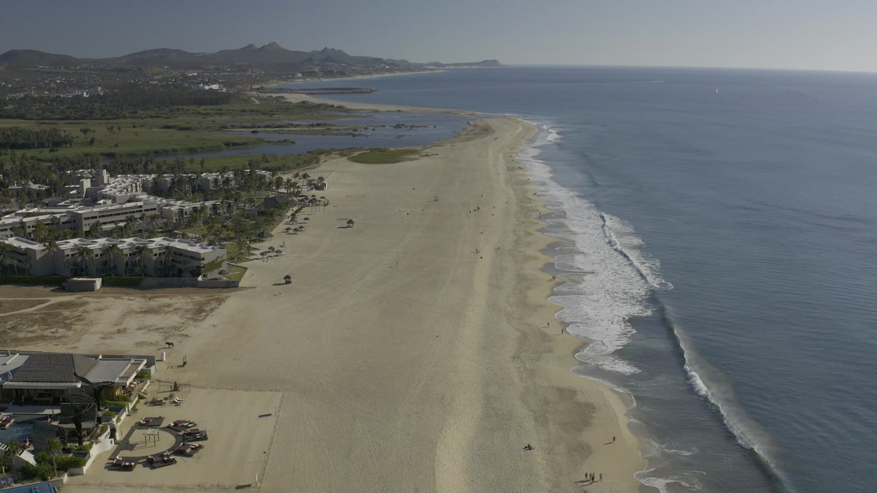 Aerial frame over Baja California Sur, Mexico showing a long sandy beach, blue ocean to the right, coastal buildings at lower left, green inland areas, distant hills, and no visible people.