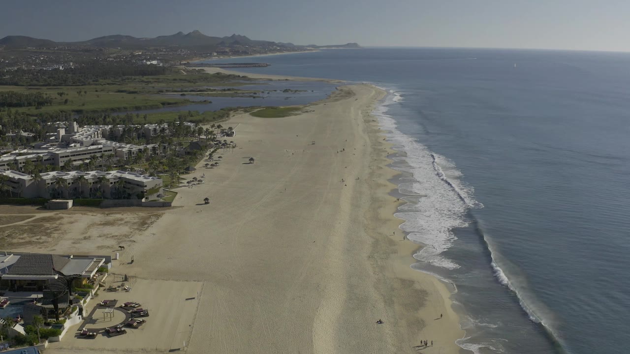 Wide drone view down a sandy shoreline with ocean waves and inland greenery.