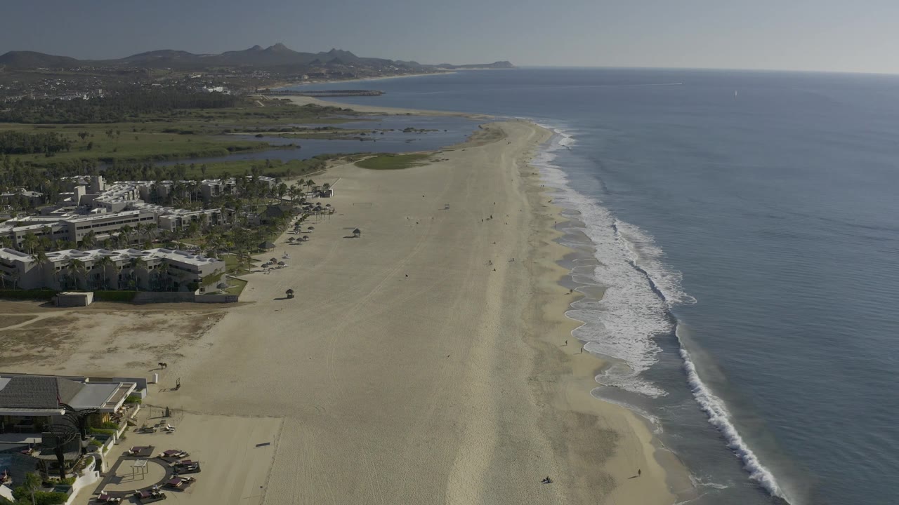Baja California Sur aerial frame with the ocean filling the right side, pale beach through the center, resort-like coastal structures at lower left, distant hills inland, and no visible people.