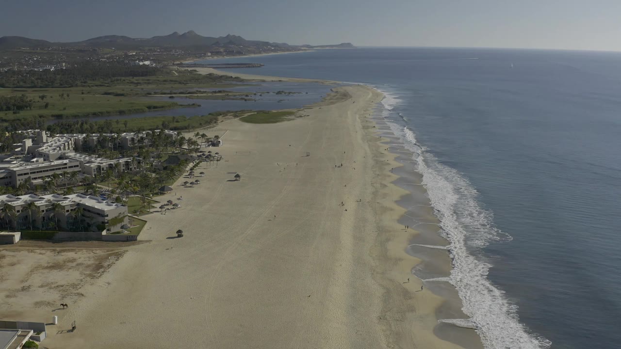 High coastal view in Baja California Sur showing a long pale beach, thin white surf lines, inland lagoon-like water, distant hills, and no visible people.