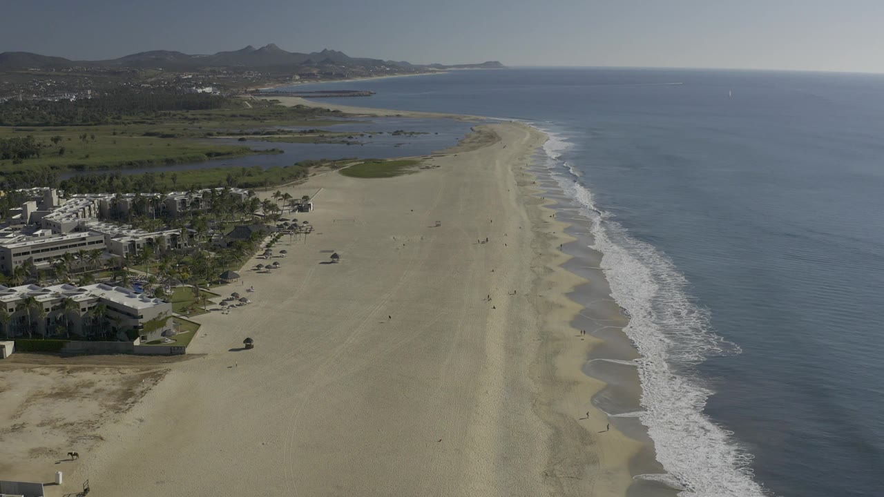Wide aerial frame over Baja California Sur with resort-edge buildings at left, a broad sandy shoreline through the center, ocean on the right, hills in the distance, and no visible people.