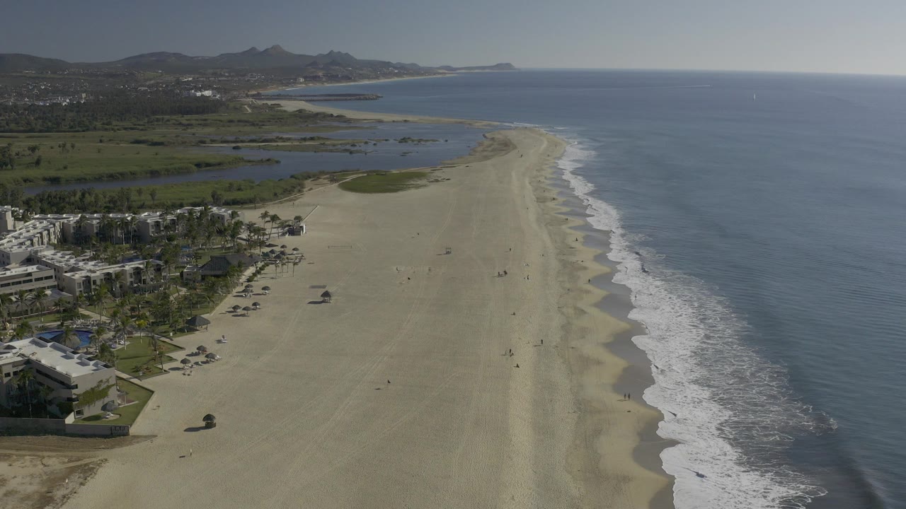 Aerial opening frame over Baja California Sur, Mexico, showing a long sandy beach beside calm blue ocean, lagoon water inland, palms and low buildings at left, distant hills, and no visible people.