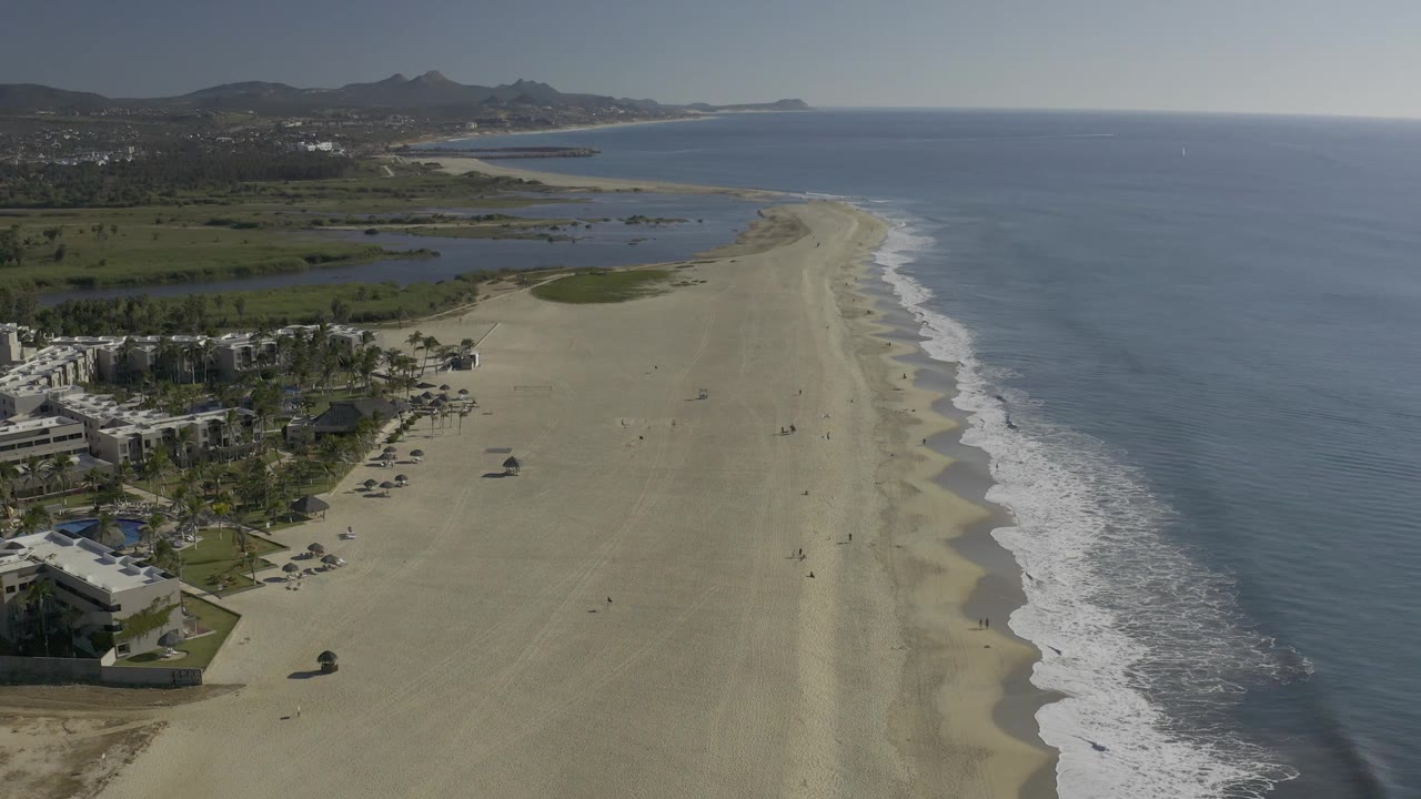 Early aerial frame over Baja California Sur coastline with the surf line running diagonally along the beach, inland lagoon water behind it, clustered palms and low buildings at left, and no visible people.