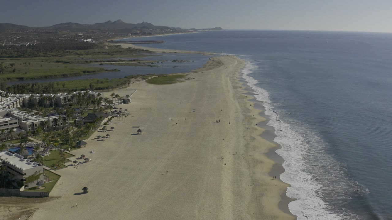 Aerial frame over the East Cape shoreline in Baja California Sur, Mexico, with more open ocean on the right, white wave bands at the shore, lagoon water inland, and no visible people.