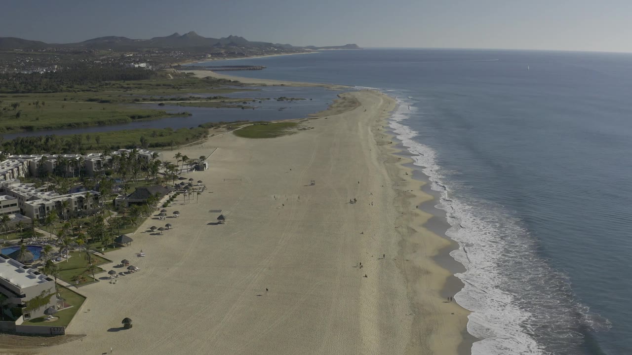 Wide aerial coastline with beach, calm sea, inland water, green vegetation, buildings, and distant ridges.