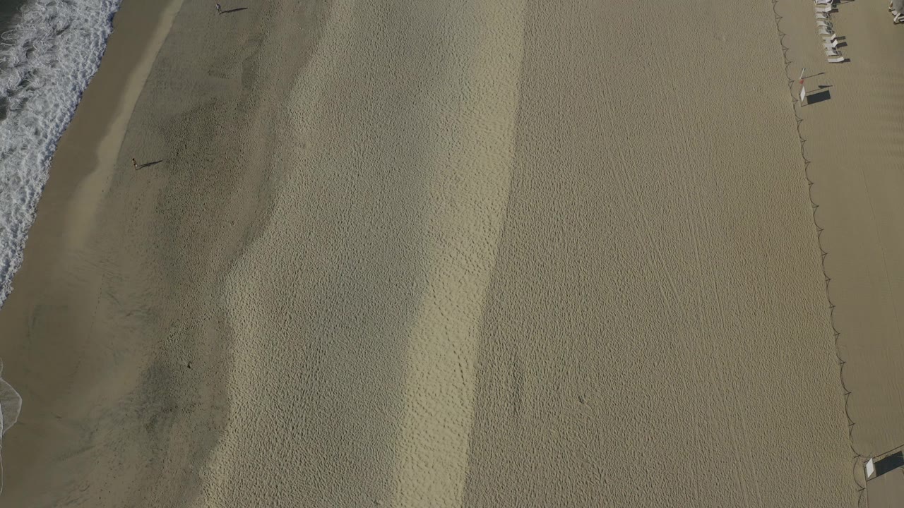 Overhead aerial view of a Baja California Sur beach with white surf at the left edge, darker wet sand near the water, and broad tan sand filling most of the frame.