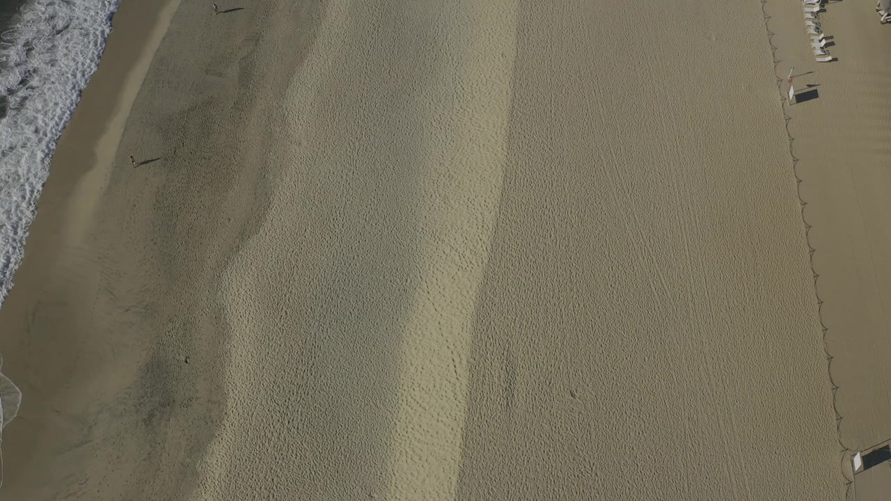 Aerial view over Baja California Sur shoreline where a white wave line borders darker wet sand and a large open section of pale beach extends to the right.