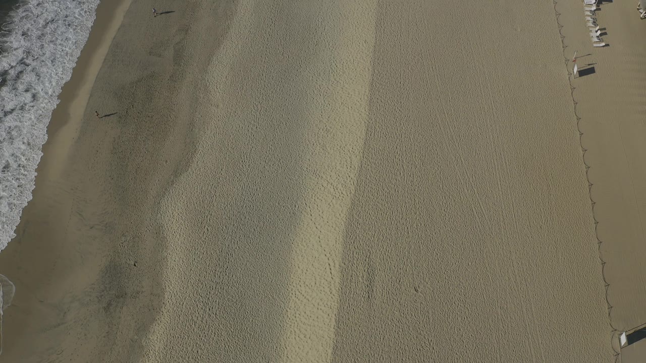 Top-down Baja California Sur beach frame with surf cropped along the left, rippled sand across the center, and small beach fixtures beginning to appear near the right edge.