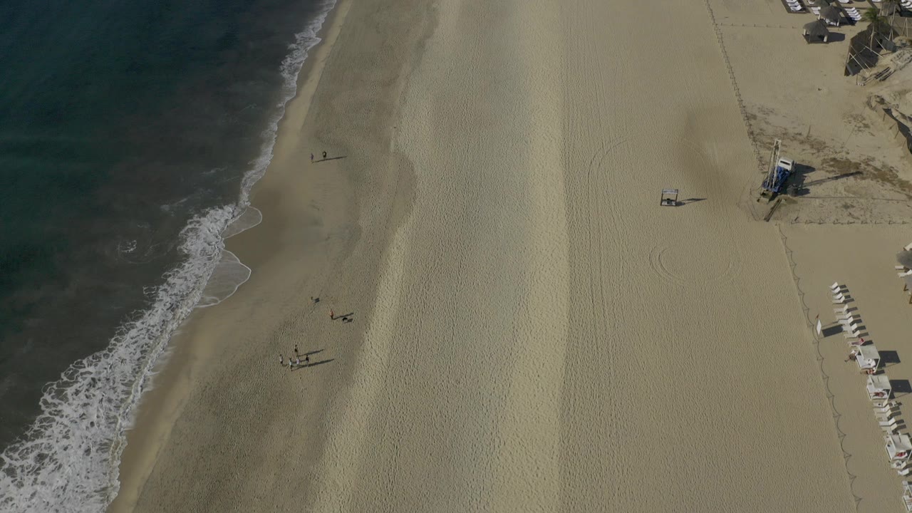Overhead view of Baja California Sur coastline with dark water on the left, pale sand across the center, a small beach structure on the right, and no visible people.