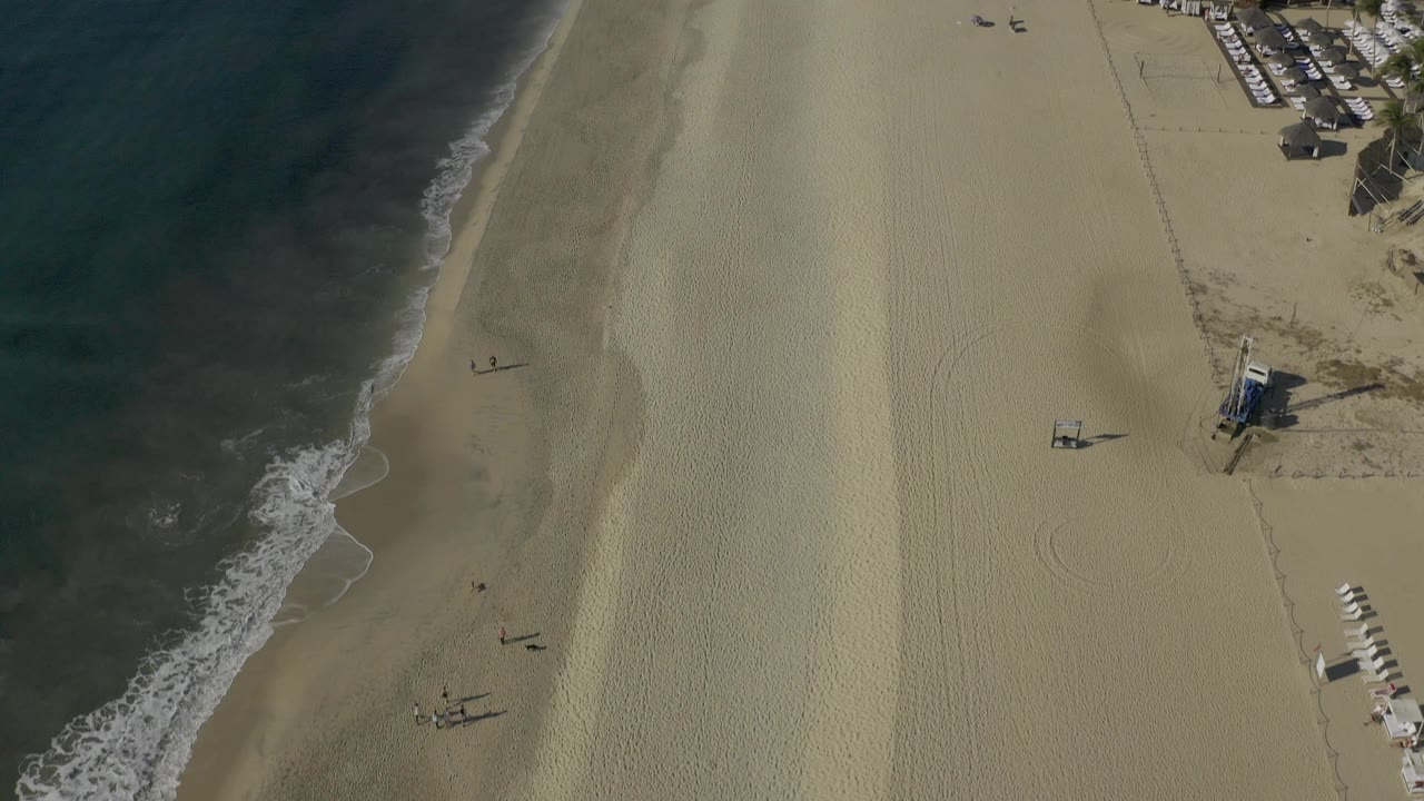 High drone view over Baja California Sur surf and sand, with a small structure near the beach and a cluster of beachfront buildings and chairs along the upper right, with no visible people.