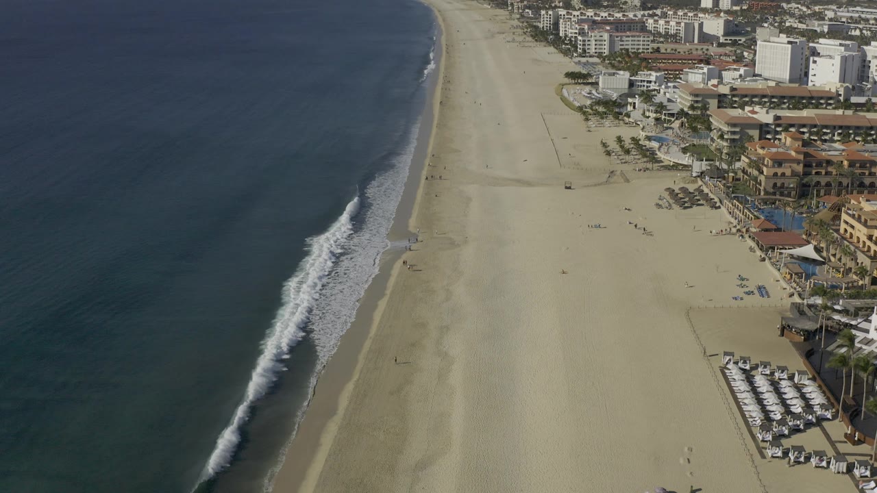 Aerial view of the Baja California Sur beach with the surf line angled down the frame, large resort buildings at right, sparse tiny figures or objects on the sand, and hazy hills beyond the coastline.