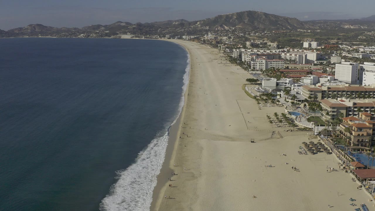 Elevated view over Baja California Sur shoreline with blue ocean on the left, tan beach down the center, coastal buildings on the right, distant hills, and no visible people.