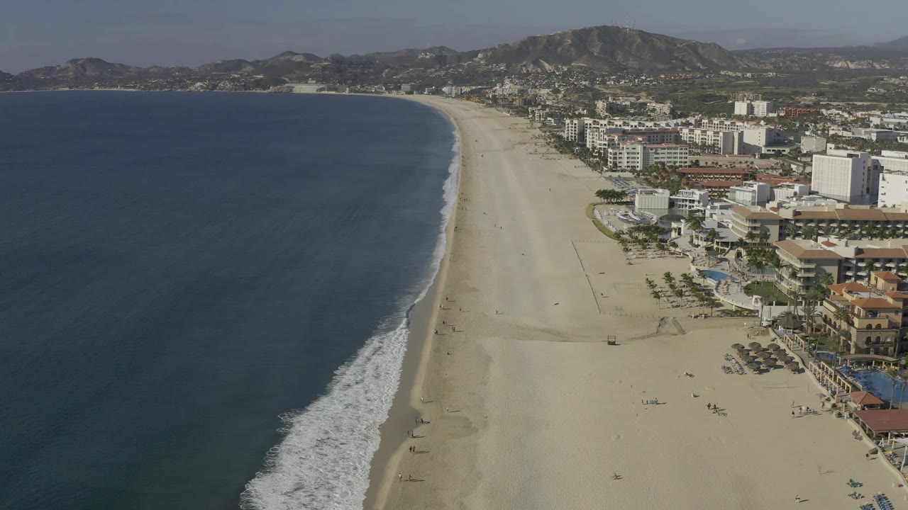 Baja California Sur beach seen from above with dark blue ocean at left, a thin surf line, pale sand, clusters of white buildings, inland hills, and no visible people.