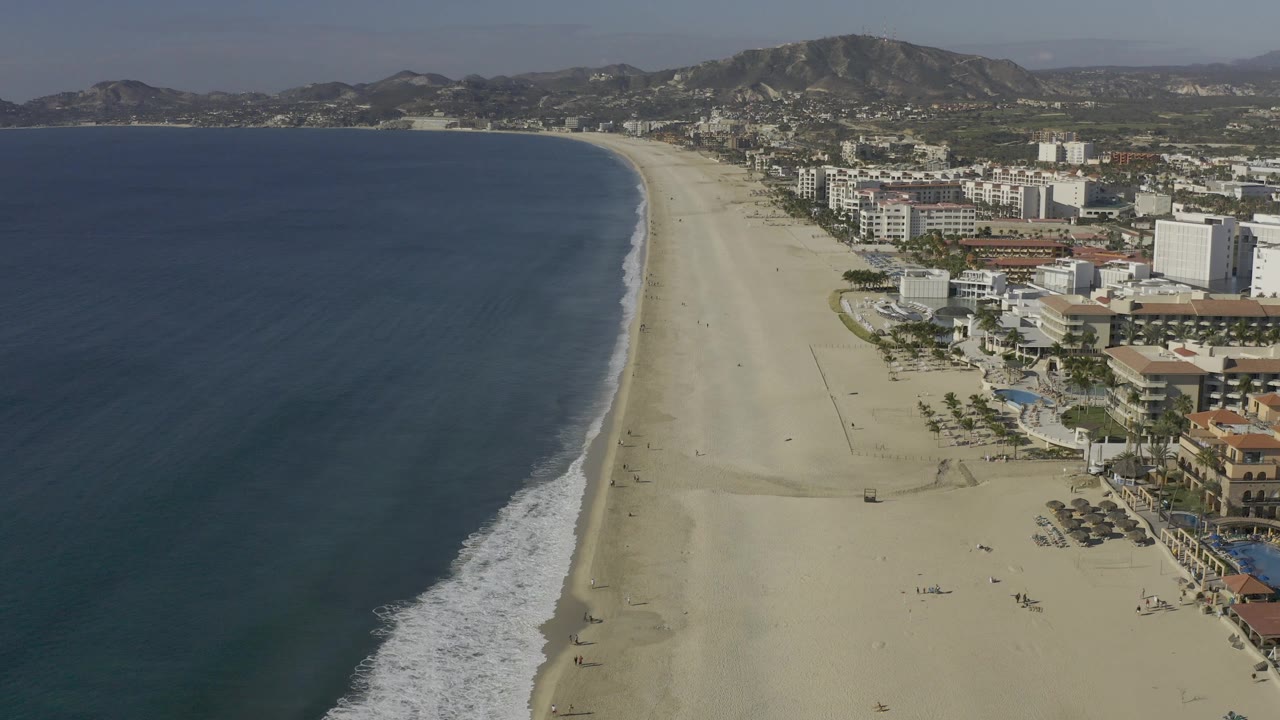 A high aerial view in Baja California Sur shows the ocean widening on the left, the beach running toward the horizon, beachfront buildings to the right, mountain terrain behind, and no visible people.