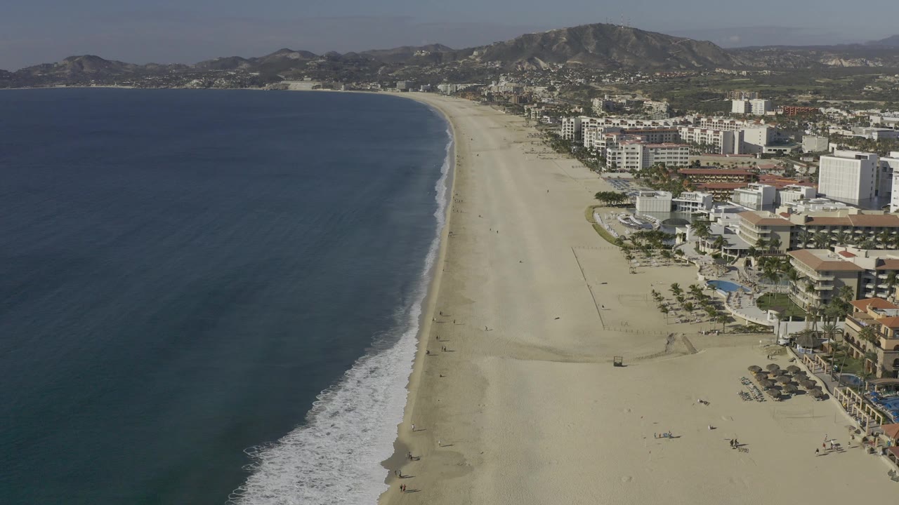 Aerial beach scene with blue sea, surf, white buildings, and a mountain ridge.