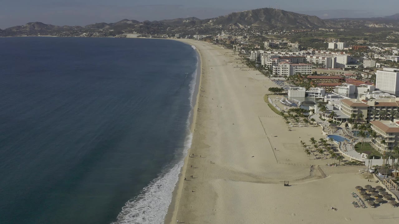 Aerial frame of Baja California Sur coastline with the beach curving into the distance, ocean surf edging the sand, hotel buildings and pools to the right, and no visible people.