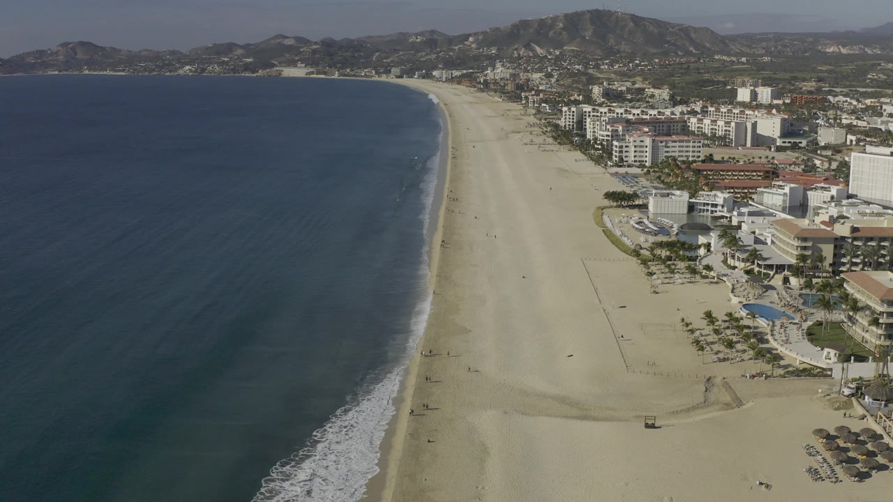 High aerial view over Baja California Sur, Mexico, showing dark ocean water, a long sandy shoreline, clusters of hotels and vacation rentals, low hills, and no visible people.