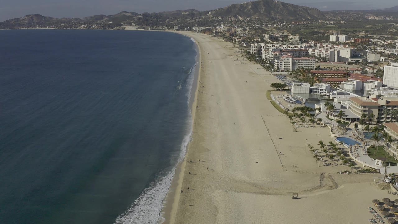 Aerial coastline frame in Baja California Sur with a darker band of water near shore, thin white surf, pale sand, hotel blocks and pool areas to the right, and no visible people.